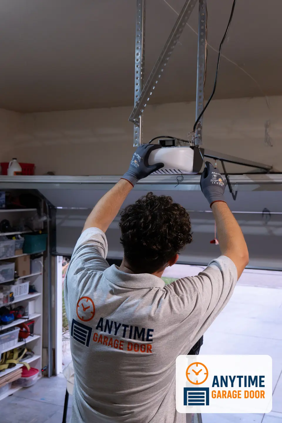 Technician in an Anytime Garage Door shirt reaches up to a garage door opener motor hanging from ceiling brackets.