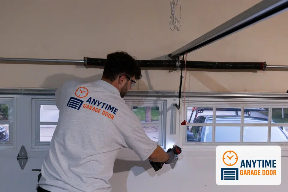 Technician uses a cordless drill on a center hinge of a garage door, with the torsion spring mounted above.