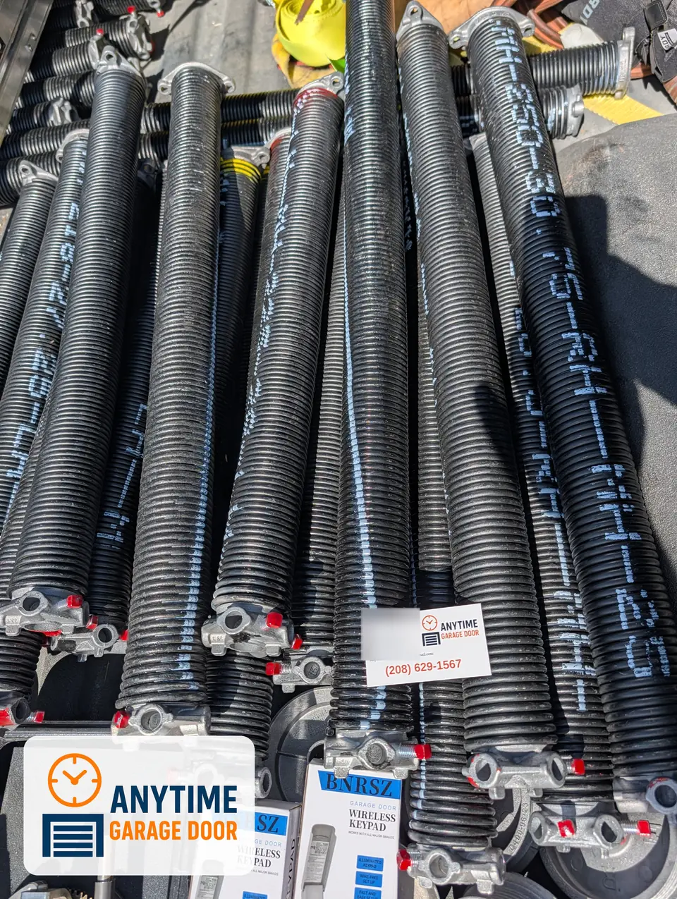 Torsion springs stacked in a truck bed with an Anytime Garage Door business card and boxed wireless keypads